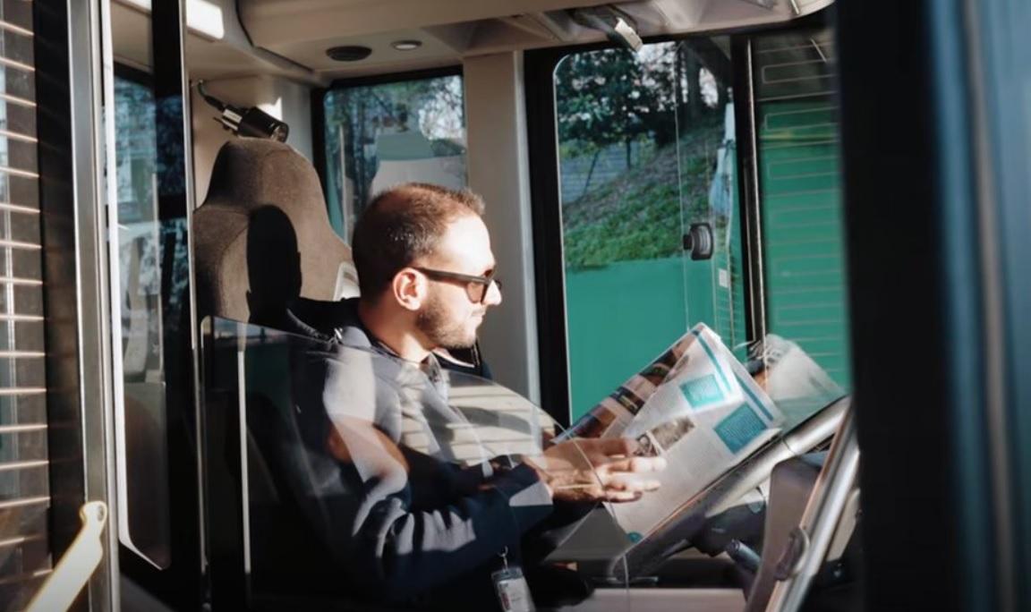 Автономен електрически автобус се движи по пътя, докато шофьорът чете вестник